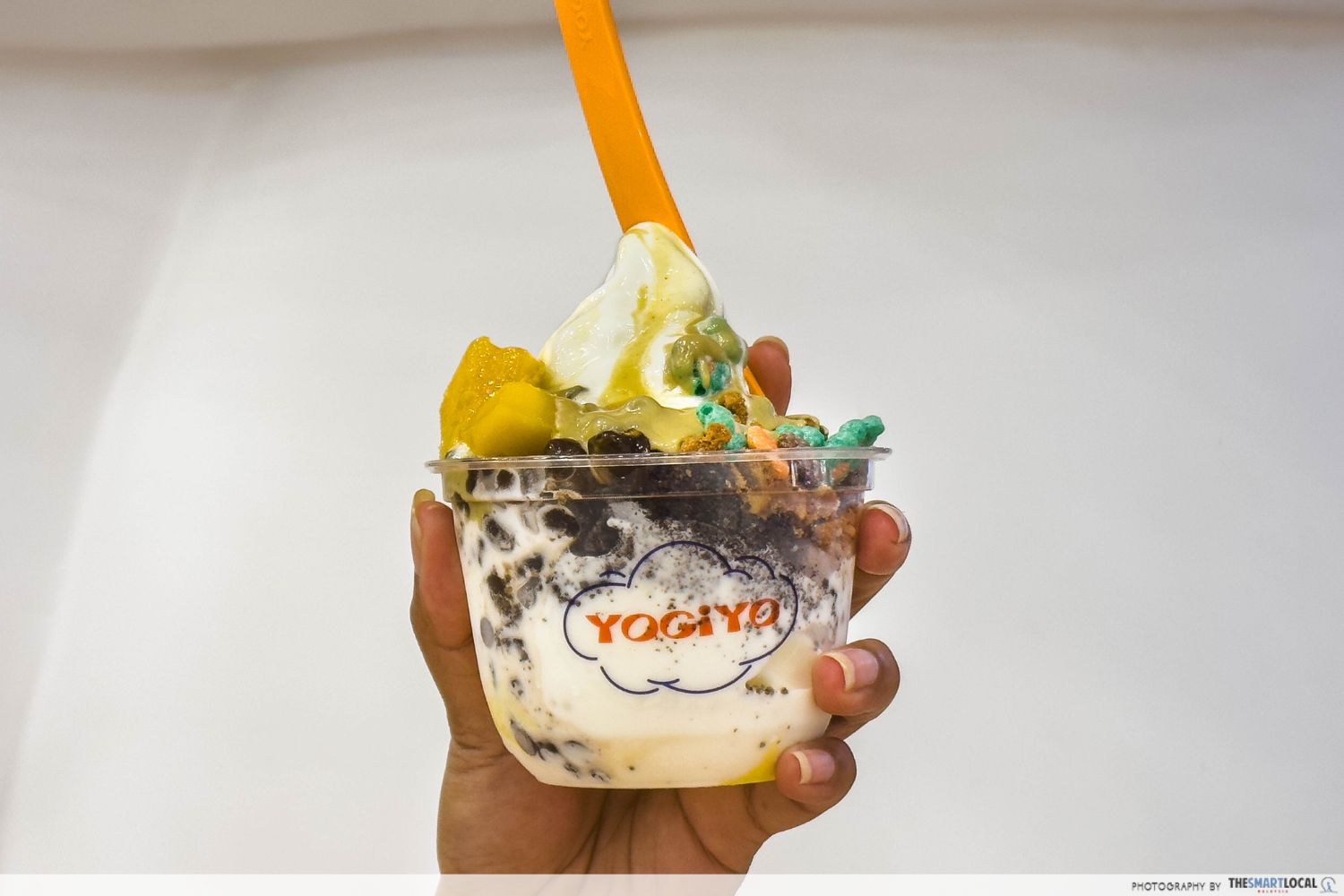 a hand holding a medium cup of froyo by yogiyo, it is topped with colourful cereals, chocolate chips, fruits, and pistachio sauce